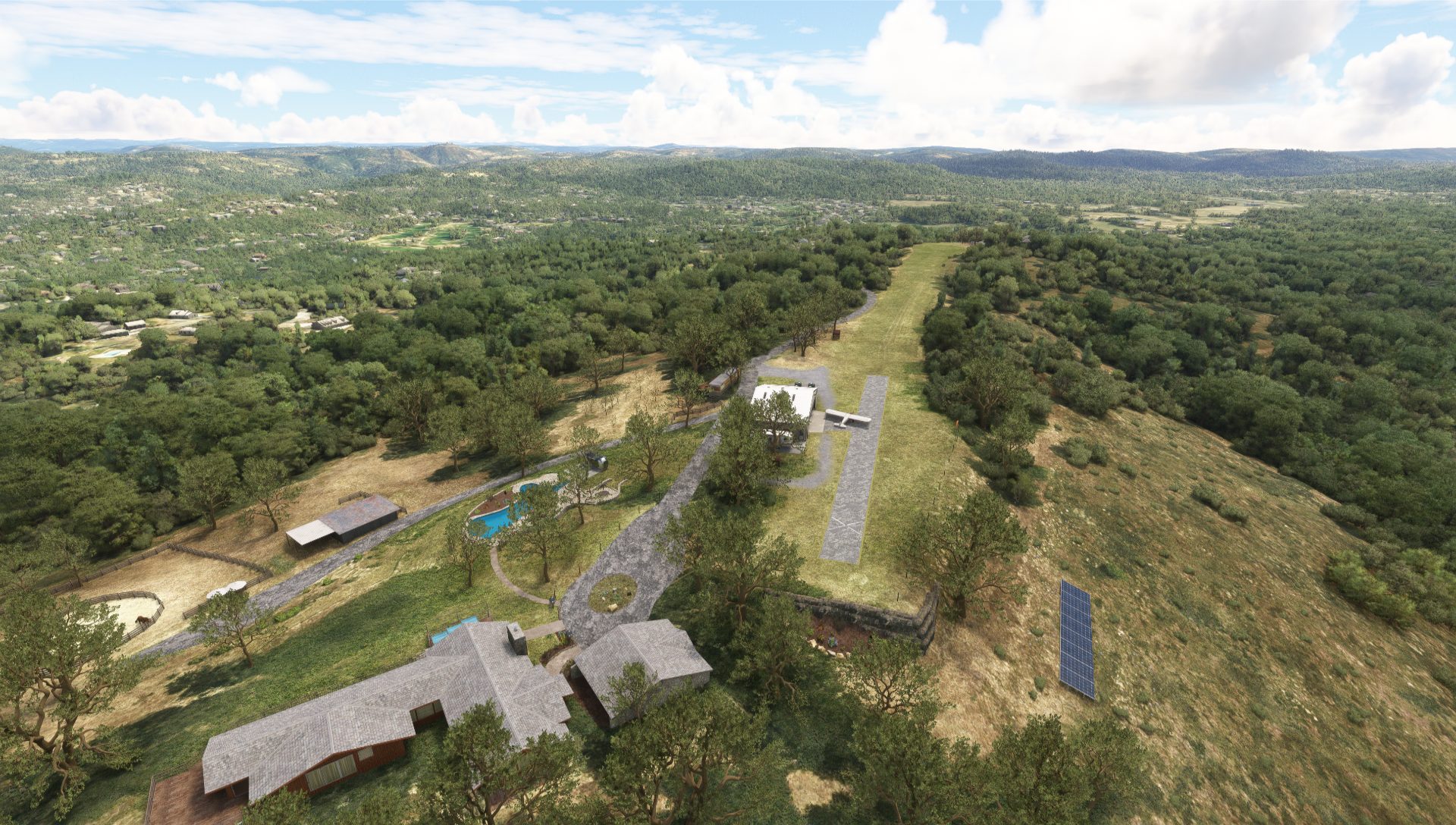 Aerial view of a rural property with buildings, a swimming pool, a small airstrip, solar panels, and surrounding hilly landscape covered in trees and grass.
