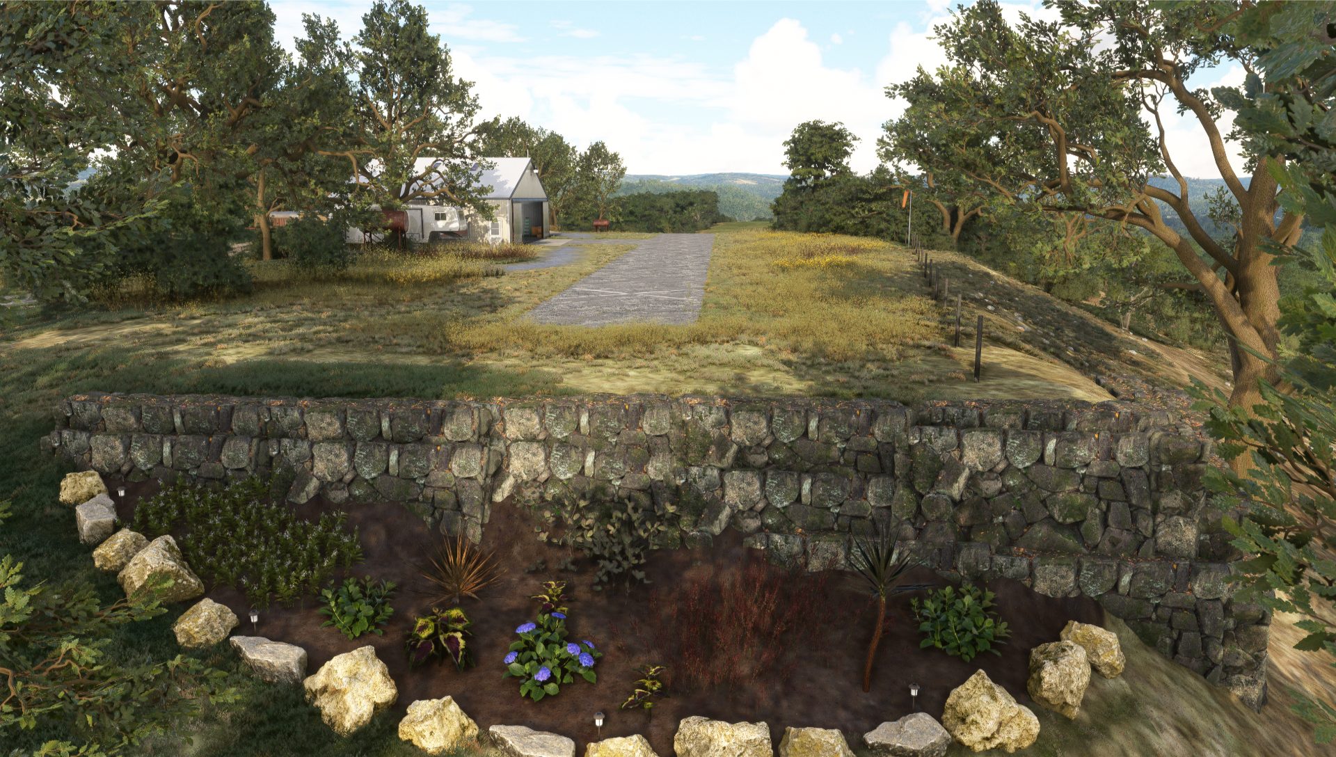 A stone retaining wall with various plants in front, grassy area above, and a hangar with a paved threshold in the background surrounded by trees.