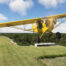 A yellow Piper Cub flies over a well maintained striped grass runway. A white, metal 3-bay Hangar is seen in the background